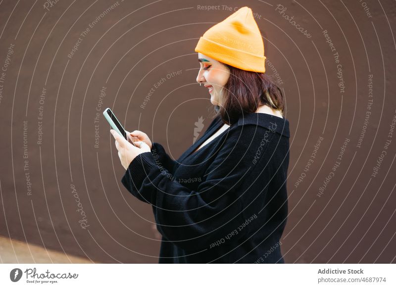 Lächelnde Frau mit Hut, die auf ihrem Smartphone surft online Browsen Textnachricht Internet informell Wand Straße Stil Funktelefon soziale Netzwerke App