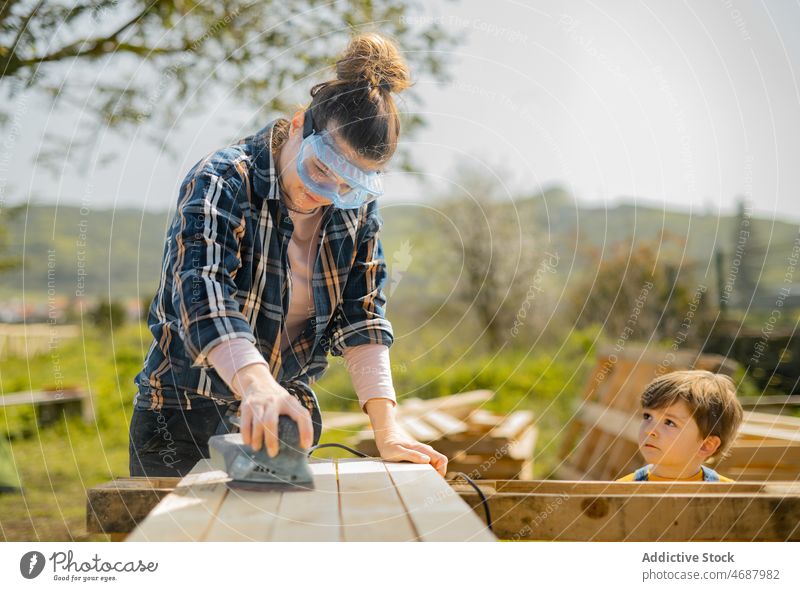 Frau schleift ein hölzernes Detail in der Nähe eines Jungen ab Schleifmaschine Instrument Werkzeug Schiffsplanken Landschaft Holzplatte Holzarbeiten Fokus