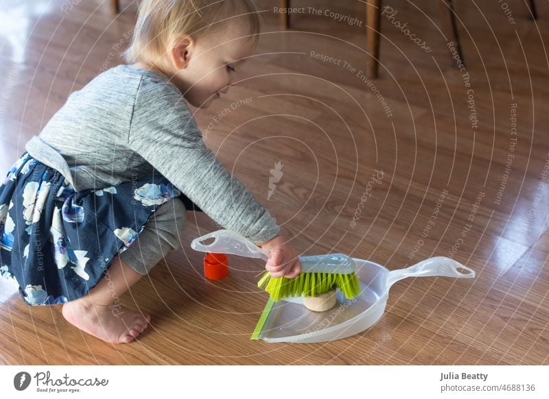 Ein 20 Monate altes Kleinkind übt das Fegen mit Kehrschaufel und Besen, um Spielzeug zu bewegen; es lernt lebenspraktische Fähigkeiten und Nachahmung Bürste