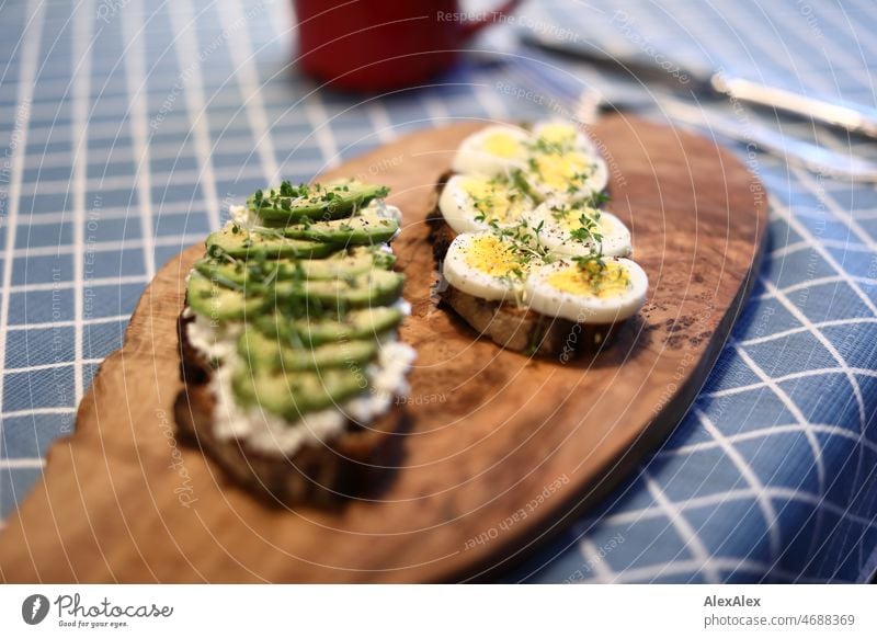 Zwei Scheiben Brot - eine mit Avocado und eine mit Ei - auf einem Olivenholzbrett auf einem Frühstückstisch gekochtes Ei Schwarzbrot Butterbrot Brett