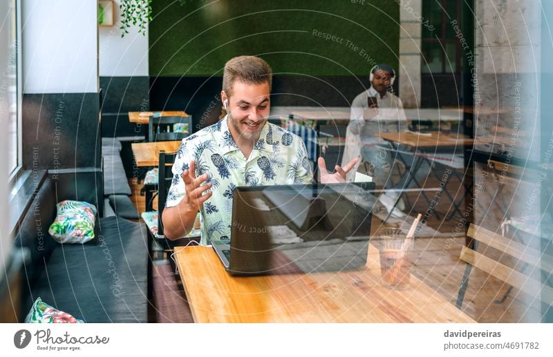 Junger Mann mit Kopfhörern und Laptop in einem Kaffeehaus Glück überrascht Erfolg gewinnen Freizeitkleidung kostenloses W-LAN Lächeln arbeiten durch das Glas