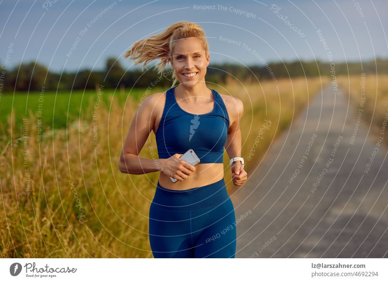 Fitte, gesunde junge Frau, die auf einer Landstraße joggt und mit einem glücklichen Lächeln voller Vitalität in einem aktiven Lebensstilkonzept an der Kamera vorbeigeht