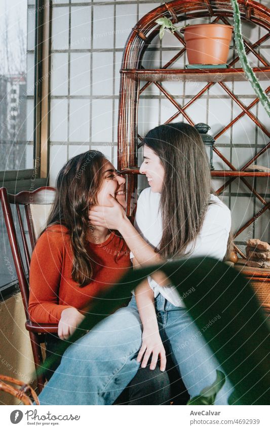 Junges und modernes lesbisches Paar kuschelt und lächelt sich gegenseitig an, während sie auf einem modernen Stuhl auf einer Galerie in einer Wohnung sitzen. Glücklich zusammen. LGBTI+ Liebe. Stolz und Recht kämpfen Konzept