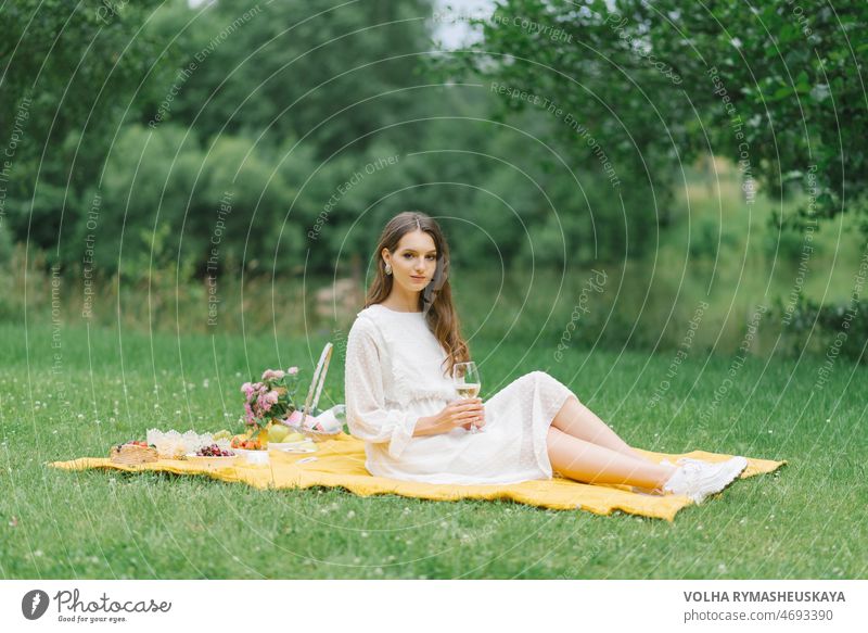 Eine schöne junge Frau in einem weißen Kleid trinkt Wein im Garten bei einem Picknick. Sommerurlaub in der Einsamkeit Korb Flasche Plaid Baguette Gesundheit