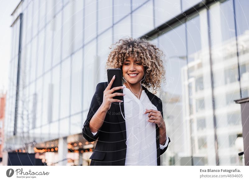 schöne lächelnde Geschäftsfrau mit Mobiltelefon und Kopfhörer in der Stadt. Gebäude Hintergrund Afro-Look hispanisch Handy Großstadt Wolkenkratzer Business jung