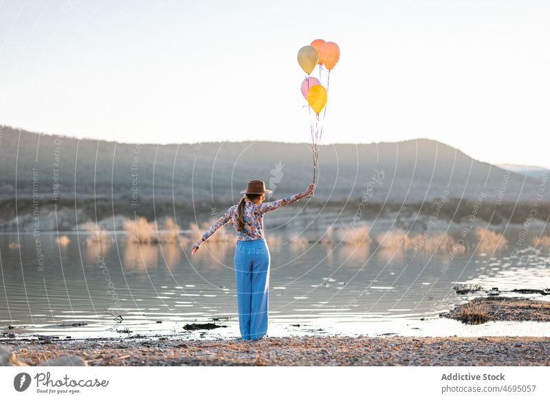Positive Frau mit Luftballons am See Ufer Wasser Landschaft Stauanlage Küste Natur Stil Hut farbenfroh Wasserseite Arme hochgezogen Sommer hell Fluss Flussufer