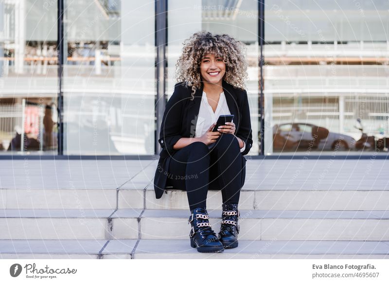 schöne lächelnde Geschäftsfrau mit Handy sitzt auf Treppen in der Stadt. Gebäude Hintergrund Afro-Look hispanisch Großstadt Wolkenkratzer Business jung