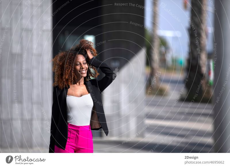 Vorderansicht einer jungen, schönen, lockigen Frau, die elegante Kleidung und eine Handtasche trägt, während sie an einem sonnigen Tag auf der Straße steht