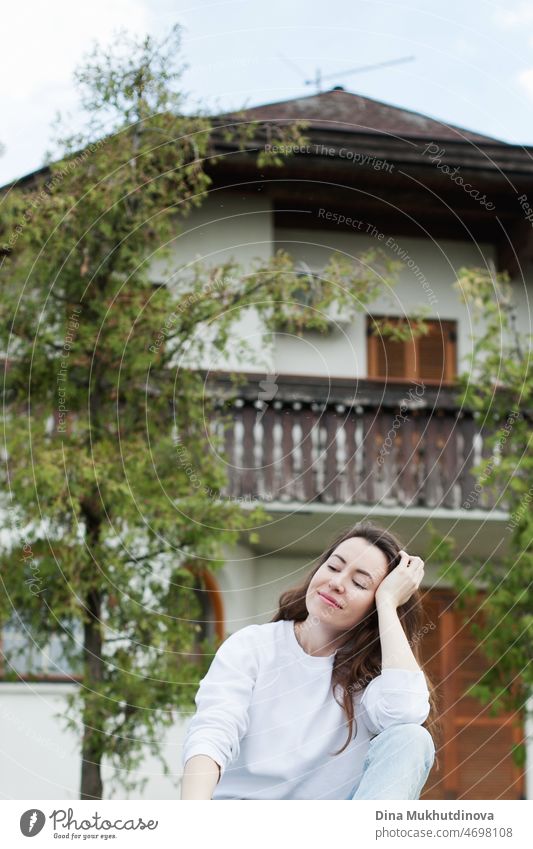 Junge Frau in weißem Sweatshirt und hellblauen Jeans sitzt in der Nähe eines Hauses auf grünem Gras im Sommer oder Frühling. Lässiges Lifestyle-Porträt. schön