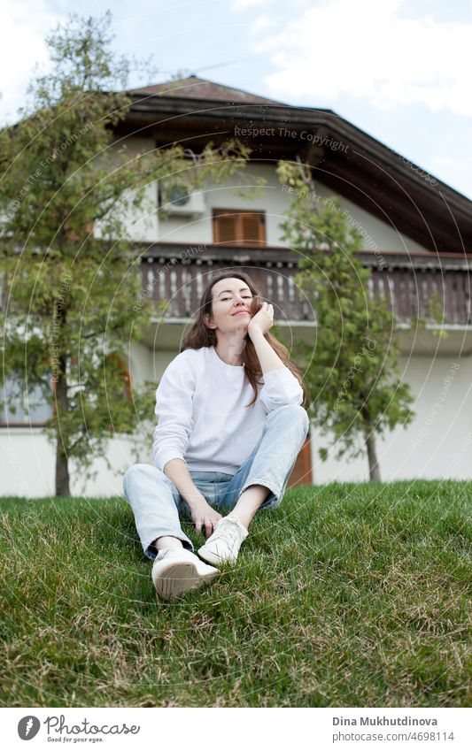 Junge Frau in weißem Sweatshirt und hellblauen Jeans sitzt in der Nähe eines Hauses auf grünem Gras im Sommer oder Frühling. Casual Lifestyle Ganzkörperporträt.