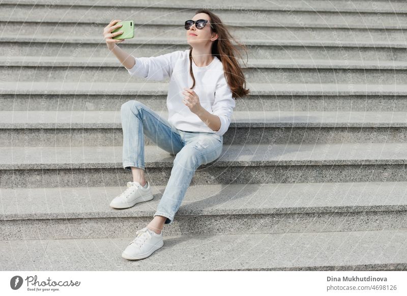 Junge Millennial-Frau in weißem Hemd, die ein Selfie macht oder mit ihrem Handy auf den Stufen eines Gebäudes videochattet. Weibliche Studentin, die auf den Treppenstufen eines Universitätsgeländes sitzt und Technologie nutzt. Echte Menschen mit Handys.