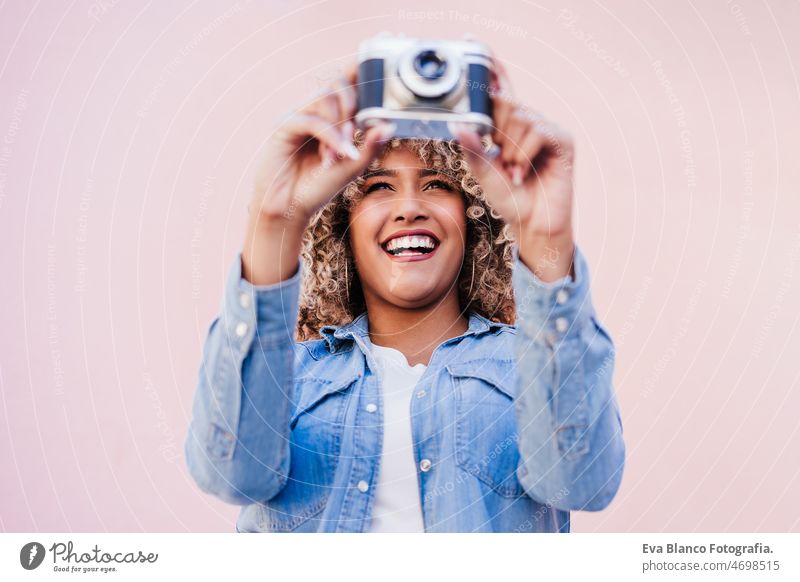 schöne glückliche hispanische Frau mit Afro-Haar hält Vintage-Kamera. rosa Hintergrund Fotokamera Fotograf Großstadt berühren Afro-Look Glück Lächeln Frühling