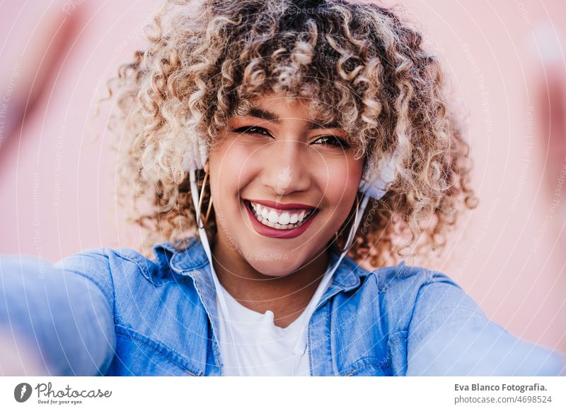 Porträt einer lächelnden hispanischen Frau mit Afro-Haar in der Stadt, die ein Mobiltelefon und ein Headset benutzt. lifestyle Selfie Handy Großstadt