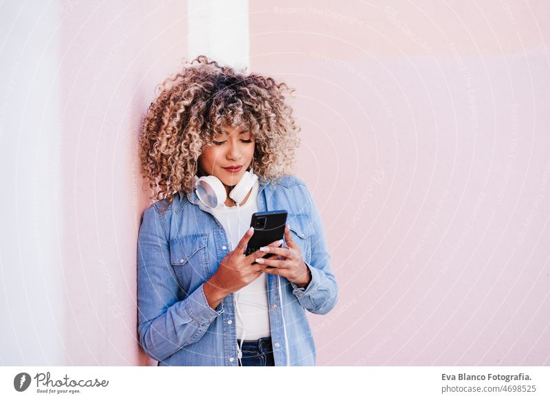 Porträt einer lächelnden hispanischen Frau mit Afro-Haar in der Stadt, die ein Mobiltelefon und ein Headset benutzt. lifestyle Selfie Handy Großstadt
