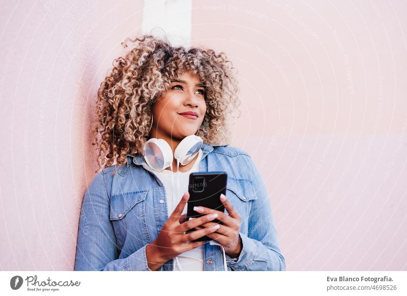 Porträt einer lächelnden hispanischen Frau mit Afro-Haar in der Stadt, die ein Mobiltelefon und ein Headset benutzt. lifestyle Handy Musik hören Großstadt