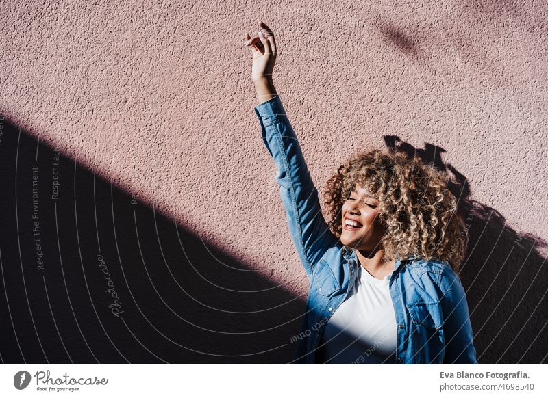 lächelnd afro hispanische Frau in sonnigen Tag mit geschlossenen Augen genießen im Freien. Stadt Lebensstil, Frühling Afro-Look Glück Lächeln Großstadt rosa
