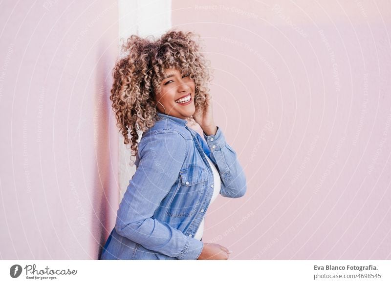 Porträt einer lächelnden hispanischen Frau mit Afro-Haar in der Stadt im Frühling. Urbaner Lebensstil Afro-Look rosa Glück Lächeln Großstadt Freizeitkleidung