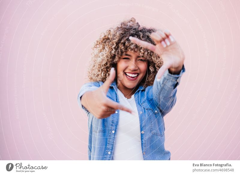 Porträt der schönen hispanischen Frau afro Haar im Freien im Frühjahr tun Rahmen mit Händen. rosa Wand Afro-Look Glück Lächeln Großstadt Frühling