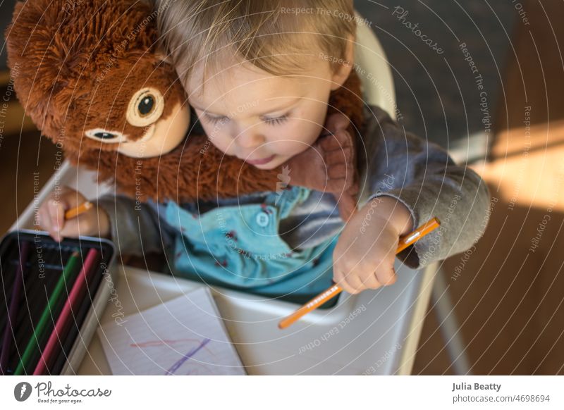Kleinkind-Mädchen, das mit einem Farbstift zeichnet, während es im Hochstuhl mit einem ausgestopften Schimpansen-Spielzeug sitzt Kind Zeichnung Bildung Schule