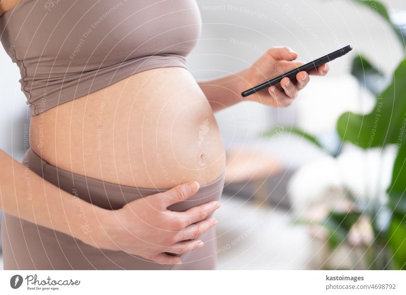 Nahaufnahme eines schwangeren Frauenbauches. Frau hält und mit Handy-Smartphone-Anwendung zu Hause Innenräume. Schwangerschaft, Technologie, Online-Shopping, Vorbereitung und Erwartung Konzept.