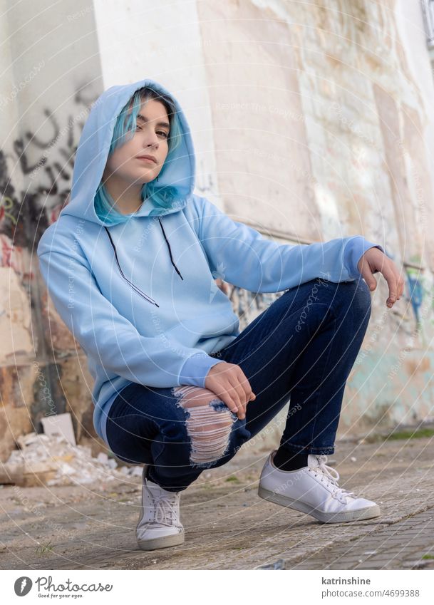 Blauhaariges Teenager-Mädchen in hellblauem Oversize-Kapuzenpulli sitzt auf ihren Hüften in der Nähe einer Graffiti-Wand hell-blau Attrappe Übergröße Jeanshose