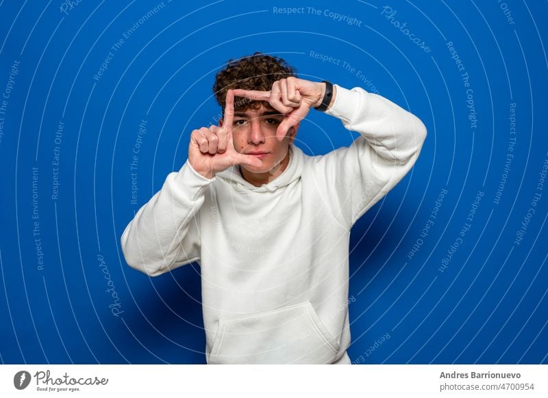 Junge gut aussehend brunette Mann mit lockigem Haar trägt weißen Sweatshirt über blauen Hintergrund lächelnd machen Rahmen mit Händen und Fingern mit glücklichen Gesicht. Konzept der Kreativität und Fotografie.