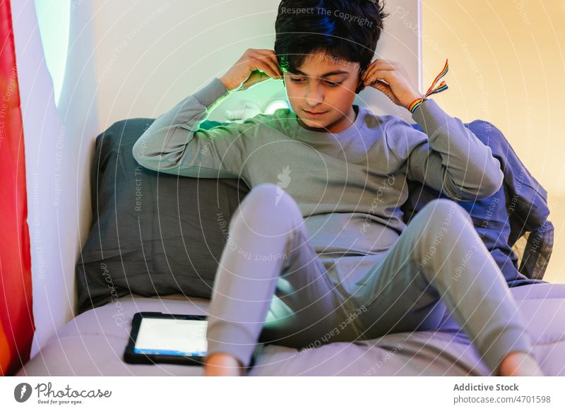 Ethnischer Jugendlicher, der Kopfhörer aufsetzt und ein Tablet benutzt Junge benutzend zuschauen Tablette Ohrstöpsel Teenager Bett nicht binär Armband Fahne
