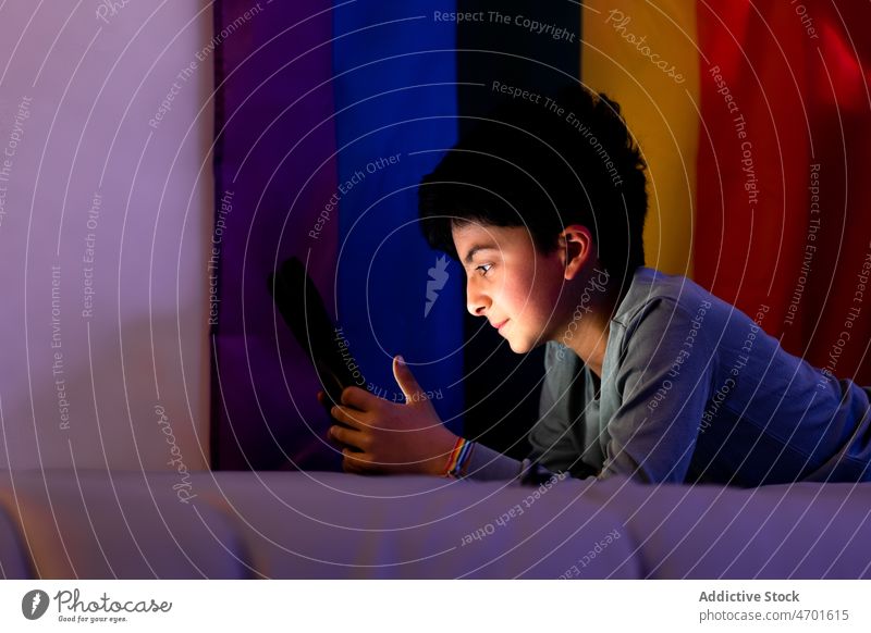 Ethnischer Junge benutzt Tablette auf dem Bett während der Schlafenszeit benutzend zuschauen online Regenbogen Fahne lgbtq Nacht nicht binär Browsen Internet