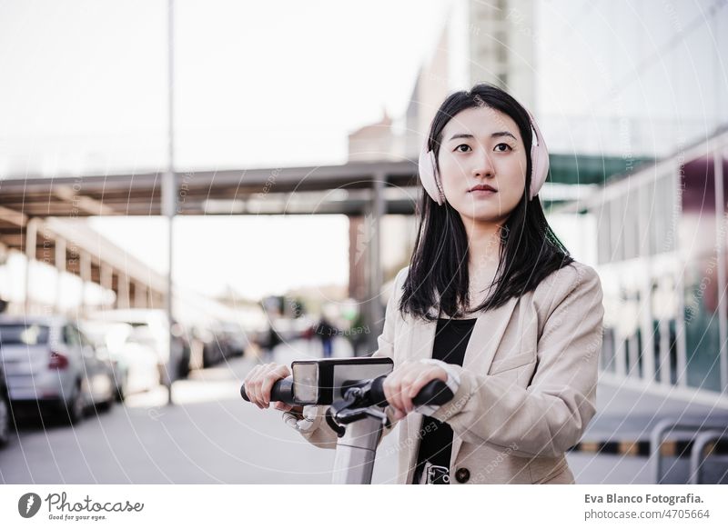 selbstbewusste Geschäftsfrau, die einen Elektroroller in der Stadt mietet. mit Headset. nachhaltiger Transport Chinesisch Frau elektrisch Tretroller Kopfhörer