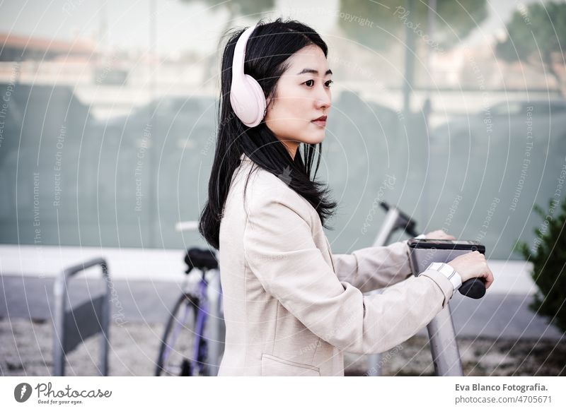 Chinesische Geschäftsfrau, die einen Elektroroller in der Stadt mietet und Kopfhörer trägt. nachhaltiger Transport Frau elektrisch Tretroller Musik Großstadt