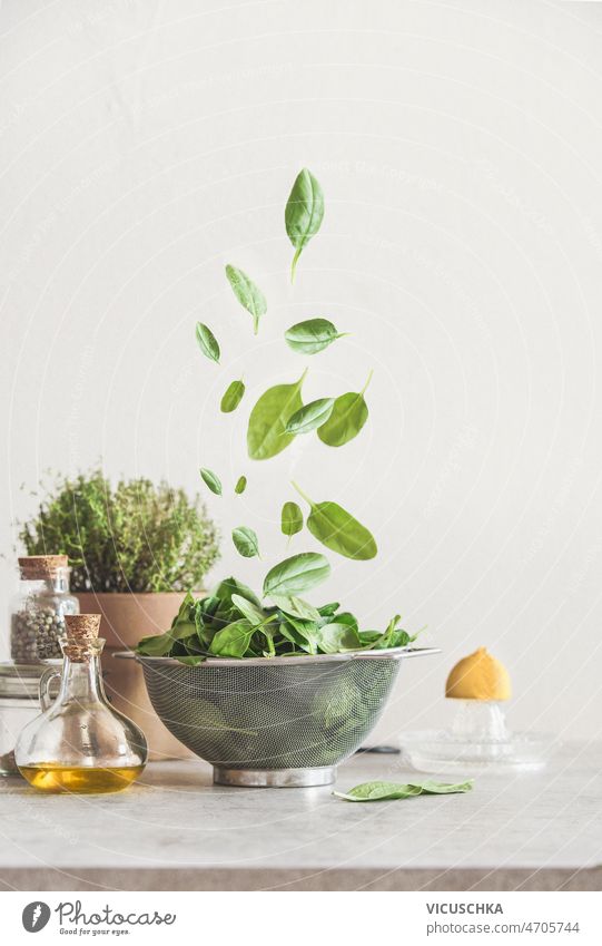 Fallende grüne Spinatblätter in einem Sieb auf dem Küchentisch mit Zutaten fallend Tisch Olivenöl eingetopft Küchenkräuter Zitrone Paprika Lebensmittel