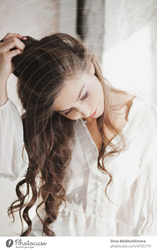 Schöne junge attraktive brünette Frau mit langen braunen Haaren in weißem Hemd, candid Lifestyle-Foto in der Nähe von Fenster mit natürlichen Morgenlicht mit romantischer Stimmung