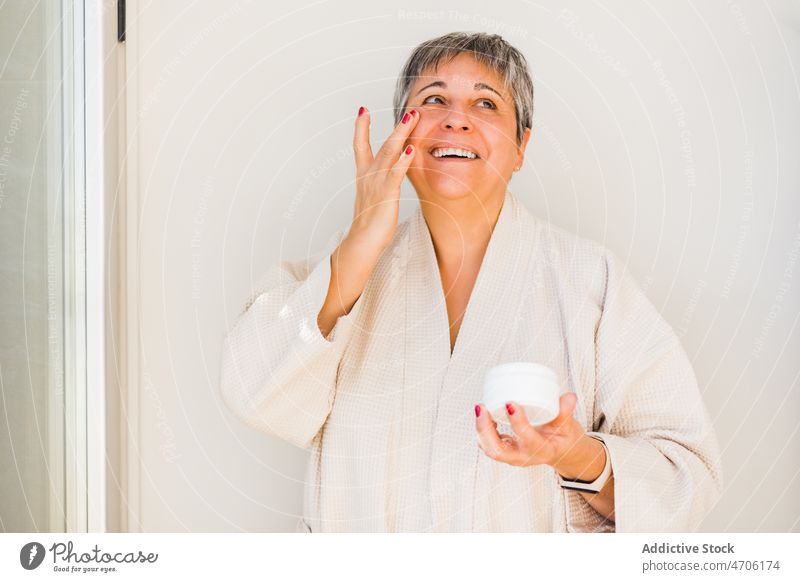 Fröhliche ältere Frau im Bademantel beim Eincremen des Gesichts Sahne bewerben Hautpflege Feuchtigkeit Kosmetik Schliere Leckerbissen körperpositiv Glück