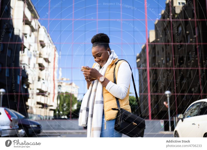 Fröhliche schwarze Frau beim Telefonieren auf der Straße Browsen zuschauend Smartphone benutzend Texten stehen herabsehend Gespräch diskutieren Kopfhörer