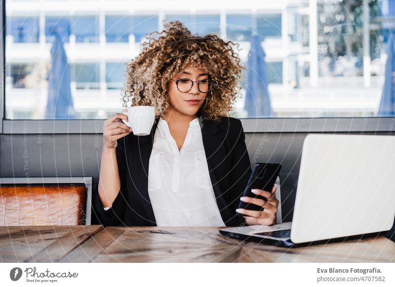 Lächelnde hispanische afroamerikanische Geschäftsfrau in einem Café, die an einem Laptop und einem Mobiltelefon arbeitet. Computer Afro-Look Frau Business