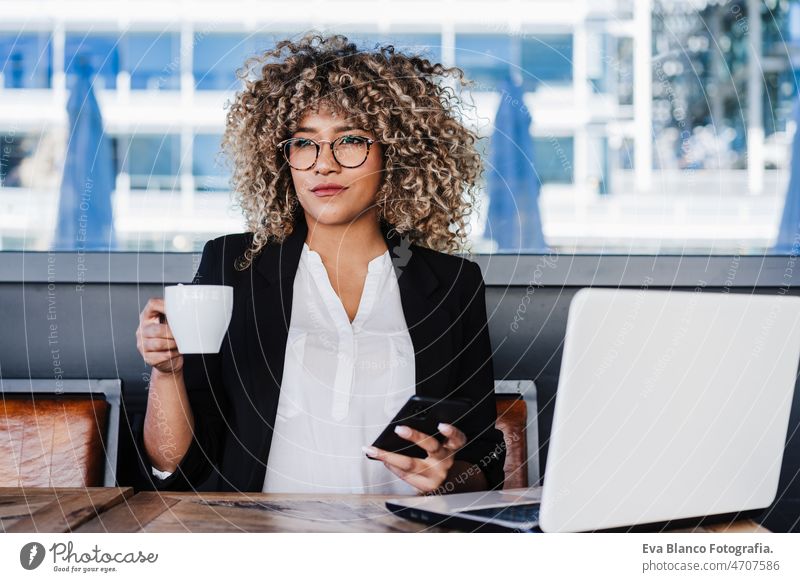 selbstbewusste hispanische afroamerikanische Geschäftsfrau in einem Café, die an einem Laptop und einem Mobiltelefon arbeitet.tech and lifestyle Computer