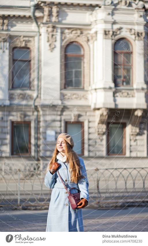 Junge schöne Frau in einem hellblauen Mantel steht auf der Straße in der Nähe von alten Gebäude in der Stadt im Sonnenlicht am sonnigen Tag. Candid Lifestyle-Porträt einer Frau, lächelnd.