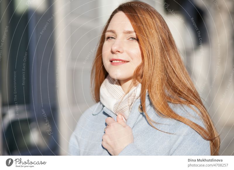 Junge schöne Frau in einem hellblauen Mantel steht auf der Straße in der Stadt im Sonnenlicht an einem sonnigen Tag. Candid Lifestyle-Porträt einer Frau, die in die Kamera schaut und lächelt.