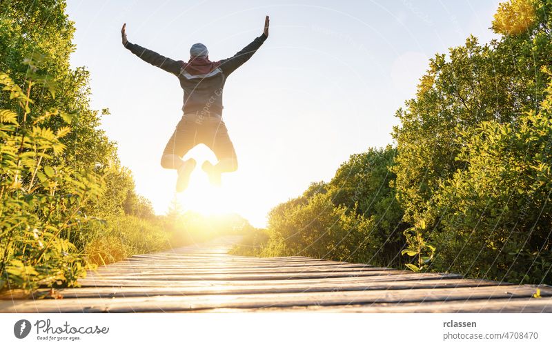 Glücklicher junger Mann, der mit erhobenen Händen und Beinen über einen Holzsteg bei Sonnenuntergang springt und das Leben genießt Anlegestelle springend frei
