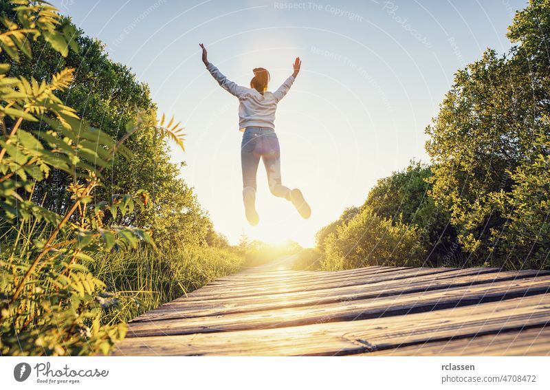 Glückliche Frau springt mit erhobenen Händen und Beinen und genießt das Leben auf einem Holzweg bei Sonnenuntergang Weg springend frei Natur Trampolin Air
