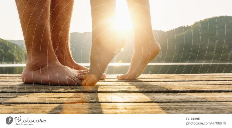 Beine auf Steg. Ein junges Liebespaar umarmt und küsst sich auf dem Steg an einem See bei Sonnenuntergang. Zwei Liebende, Mann und Frau barfuß in der Nähe des Wassers auf einem Steg. Glückliche Liebe Momente zusammen.