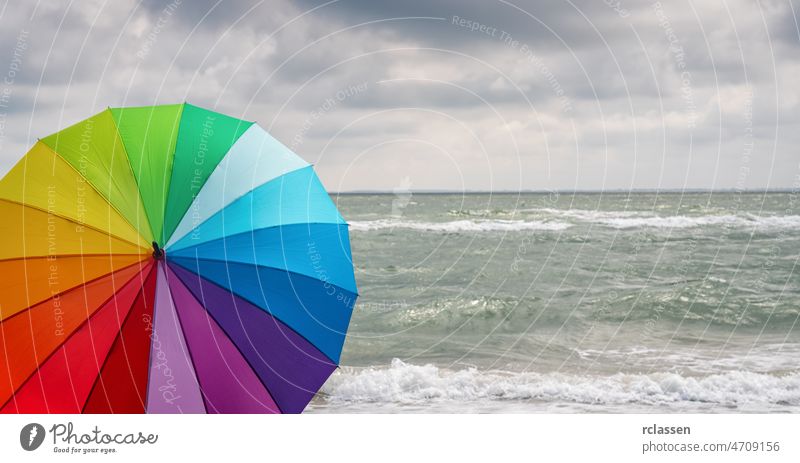Bunt von Regenschirm am Strand und Schaum der Meereswellen bei einem Gewitter, Wetter Konzeptbild farbenfroh Regenbogen Cloud Unwetter Textfreiraum Europa