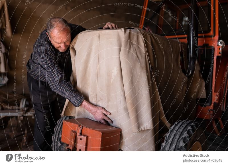 Älterer Mechaniker, der in der Garage einen Traktor mit einem Tuch abdeckt Mann Arbeiter Gewebe Bauernhof Dienst alt Reparatur männlich ländlich Wehen Job Beruf