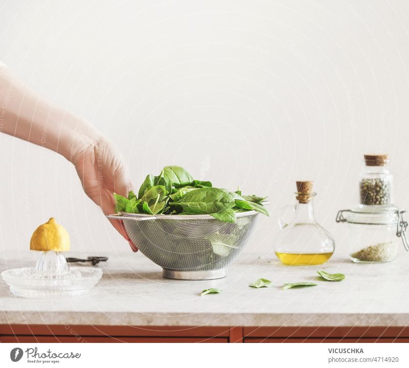 Frau hält ein Metallsieb mit rohen grünen Spinatblättern auf dem Küchentisch in der Hand Beteiligung Sieb gefüllt Blätter Tisch Zitrone Olivenöl Kräutersalz