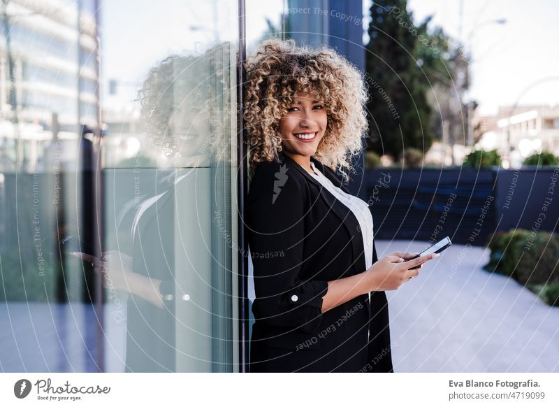 schöne lächelnde Geschäftsfrau mit Mobiltelefon und Kopfhörer in der Stadt. Gebäude Hintergrund Afro-Look hispanisch Handy Großstadt Wolkenkratzer Business jung