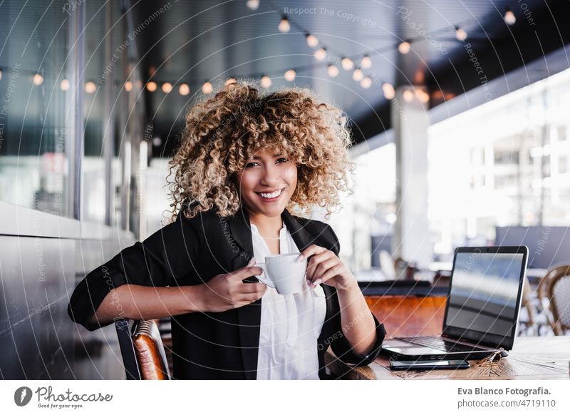 Selbstbewusste hispanische Geschäftsfrau in einem Café, die an einem Laptop und einem Mobiltelefon arbeitet. Technik und Lifestyle Computer Afro-Look Frau