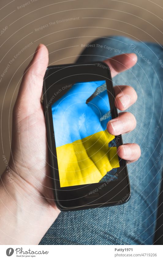 Nahaufnahme einer Hand die ein Smartphone hält, auf dessen Display die Hand, bemalt in ukrainischen Nationalfarben,durchscheinend zu sehen ist, die es hält