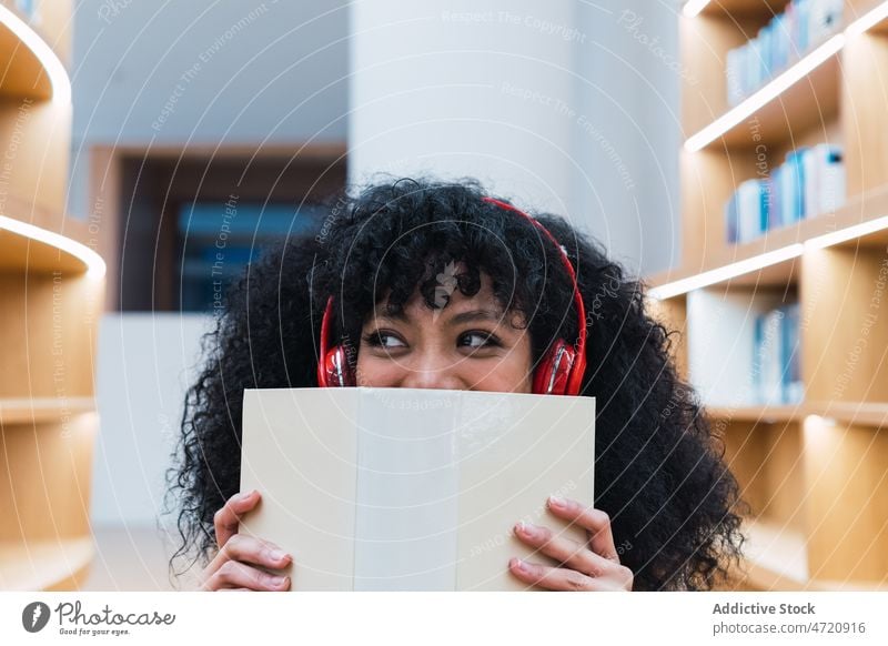 Positive ethnische Dame mit Kopfhörern und aufgeschlagenem Buch in der Bibliothek Frau meloman Musik verträumt zuhören interessant Literatur träumen Regal