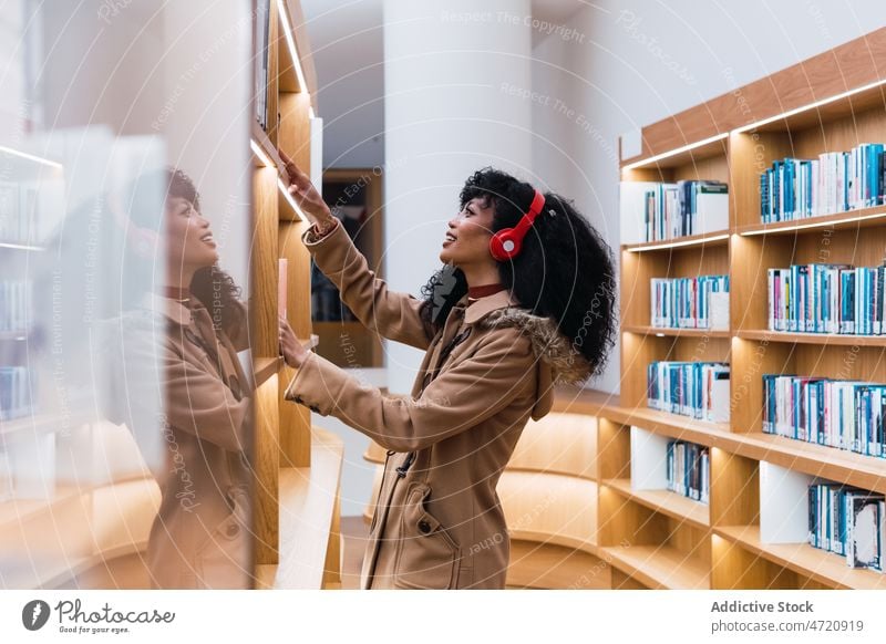Zufriedene Frau mit Kopfhörern, die ein Buch in der Bibliothek liest melomaniac Regal Schüler Bücherschrank positiv wählen Bücherregal Lächeln Musik zuhören