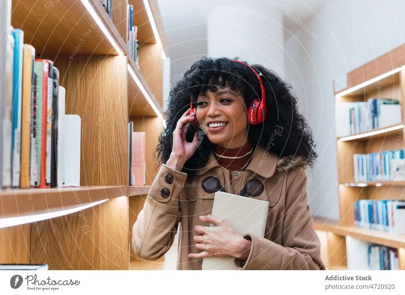 Positiver schwarzer weiblicher Meloman, der in einer Bibliothek über sein Smartphone spricht Frau Telefon benutzend sprechen meloman Regal Buch Kopfhörer